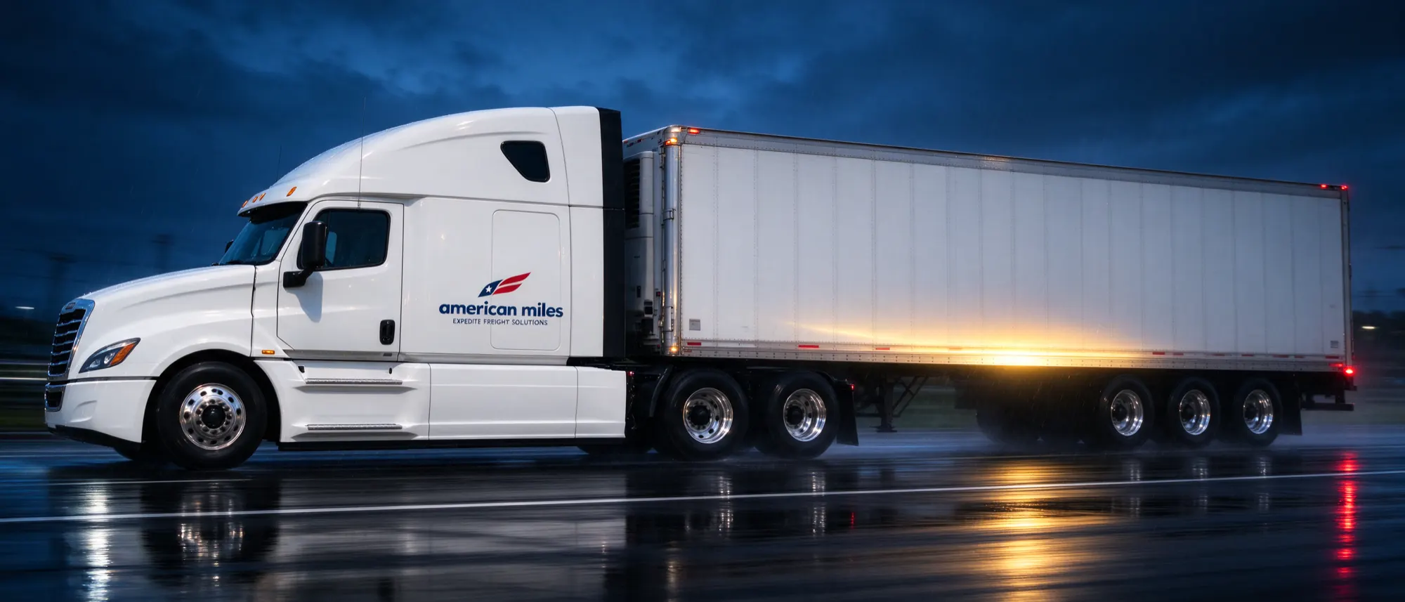 American Miles Freightliner Cascadia sleeper tractor pulling a branded 53-ft dry van on a wet interstate at dusk